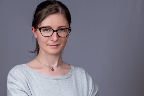 Young professional woman headshot wearing glasses on a grey background with hair tied back