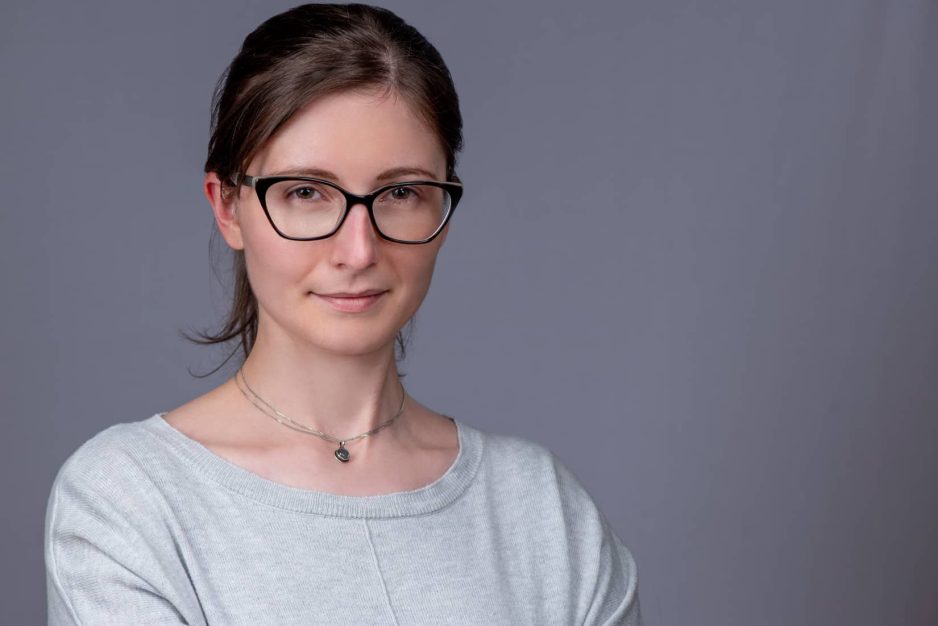 Young professional woman headshot wearing glasses on a grey background with hair tied back