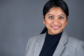Headshot of a professional woman with a black top and grey jacket on a grey background