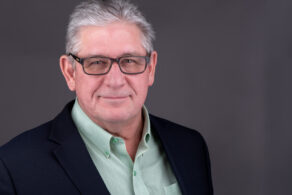 Warm looking smile headshot of an older man with a black jacket green shirt on a dark background