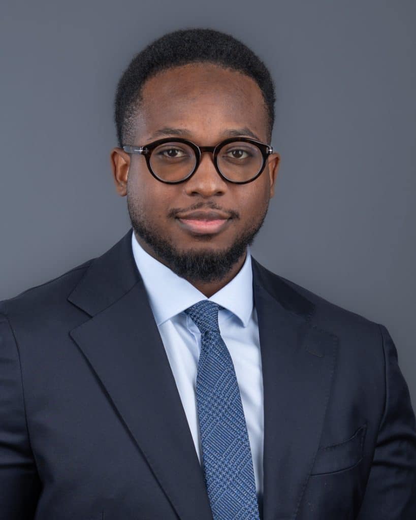 Professional headshot of young man wearing round glasses blue suit against darker grey background