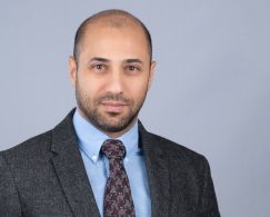 Young professional headshot of a younger man on a grey background in a tweed jacket with a button down collar and tie.